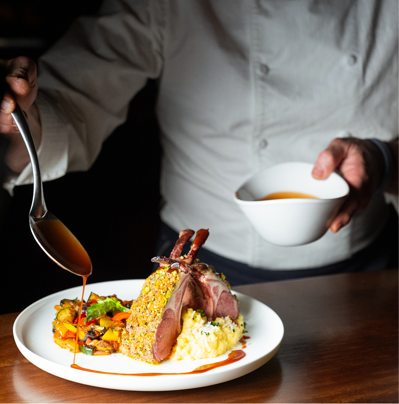 Chef drizzling sauce over a plated rack of lamb served with creamy risotto and colorful roasted vegetables.