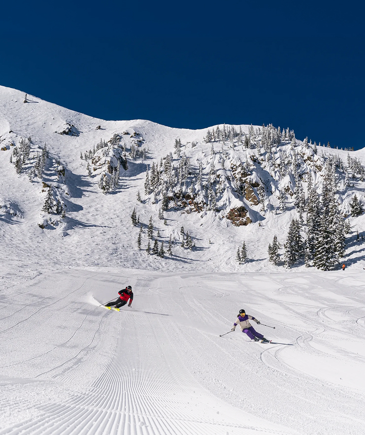 Two skiers carve down a snow-covered mountain slope at Taos Ski Valley.